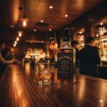重厚な木のカウンターに置かれたジャックダニエルのボトルとロックグラス。暖色の照明が琥珀色の液体を照らし、大人の隠れ家のような洗練された雰囲気を演出することで「ダサい」というイメージを払拭するビジュアル